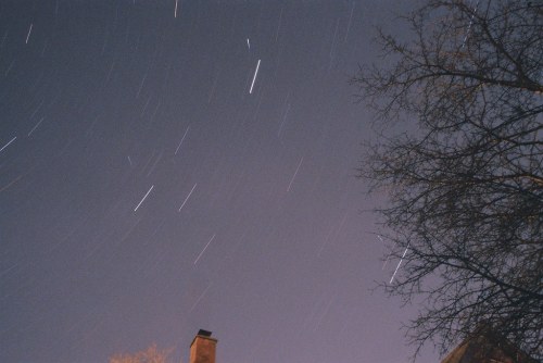 Star trails in a night sky, with a silhouette of a tree and a chimney in the foreground.
