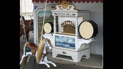 A vintage carousel organ with a white horse in motion, featuring decorative elements and a scenic painting.