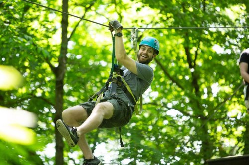 A person in a blue helmet zip-lining through a lush green forest, smiling with excitement.
