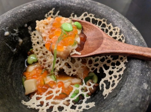 A wooden spoon holds a bite of food with vibrant toppings, resting in a textured bowl with a lace-like garnish.