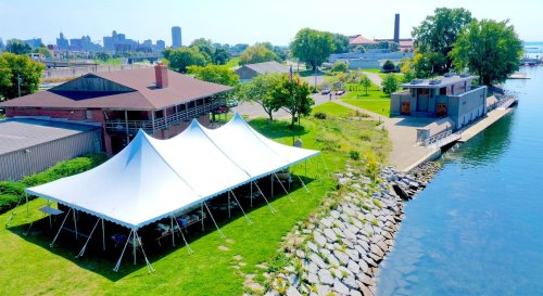 A large white tent on green grass by the water, with a city skyline in the background and a modern building nearby.