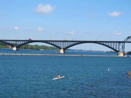 A person rows a boat on a calm river beneath a large arch bridge on a sunny day.