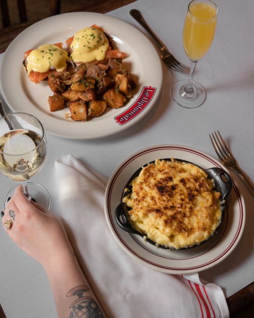 A hand holds a wine glass beside a plate of mac and cheese, a plate of eggs benedict, and a mimosa on a table.