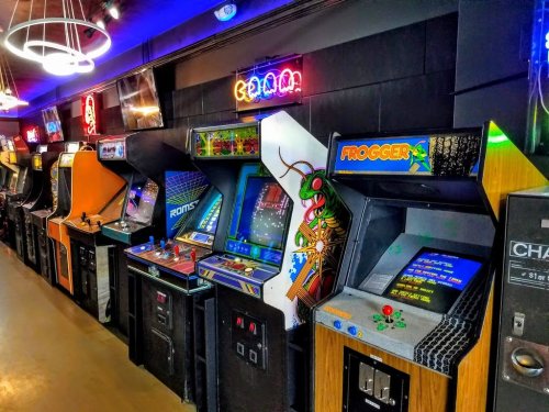 A row of vintage arcade machines, including "Frogger," with colorful neon lights in a retro gaming space.