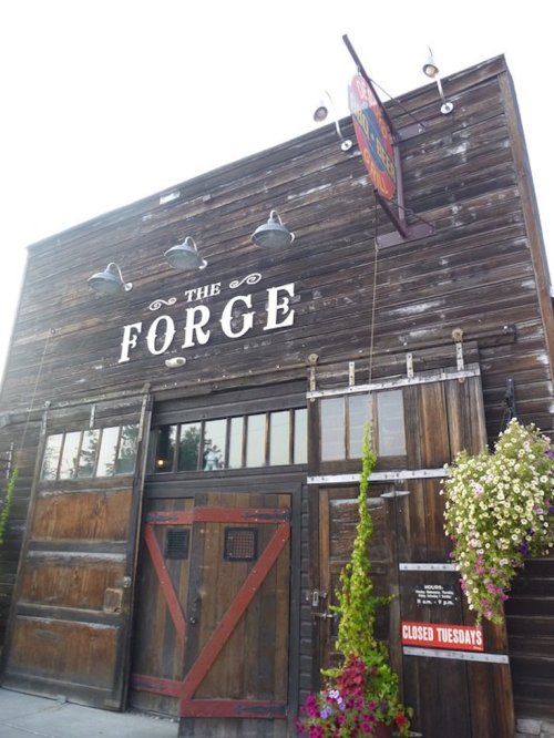 Exterior of a rustic building with a sign reading "The Forge," featuring wooden doors and hanging flower baskets.