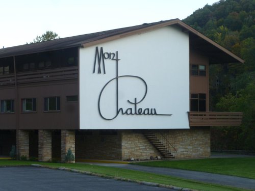 A modern building with a large sign reading "Mont Chateau" on a white wall, surrounded by greenery and hills.