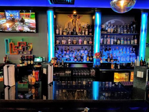 A vibrant bar with colorful lighting, shelves of liquor, and a TV displaying a sports program. Decor includes playful figures.