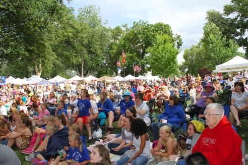 A large crowd gathers in a park for an outdoor event, with tents and trees in the background. Families and children are present.