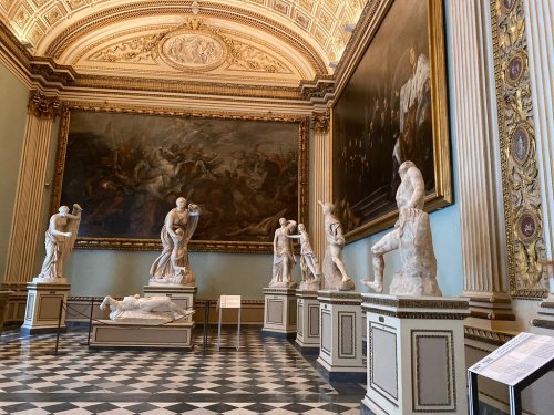 A grand museum interior featuring classical sculptures and a large painting on the wall, with intricate architectural details.