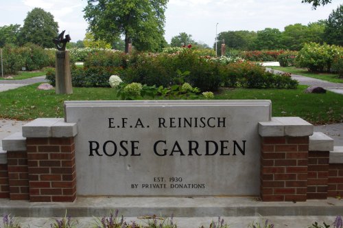 Sign for E.F.A. Reinisch Rose Garden, established in 1930, surrounded by greenery and flowers.