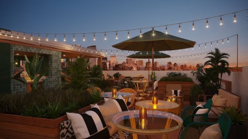 Rooftop lounge with string lights, plants, and seating, overlooking a city skyline at sunset.