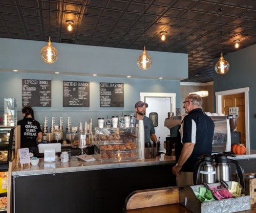 A cozy coffee shop with a counter, barista, and customers. Pastries are displayed, and menu boards are visible.
