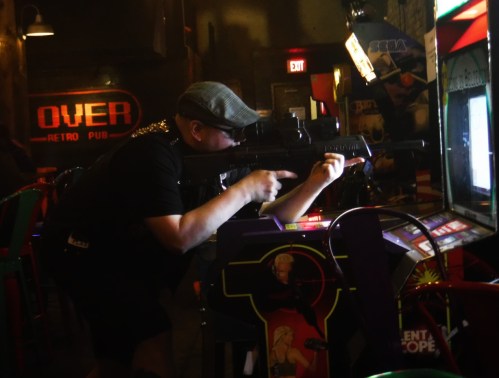 A person aims a toy gun at an arcade game in a retro pub, with dim lighting and colorful decor in the background.