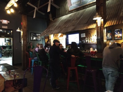 A lively bar scene with patrons at the counter, colorful stools, and a rustic decor featuring a metal roof.