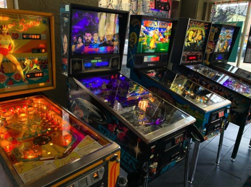 A row of colorful pinball machines with various themes and illuminated playfields in a lively arcade setting.