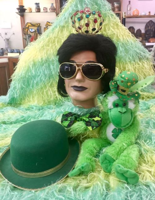 A mannequin with sunglasses and a crown, surrounded by green decor, a green hat, and a plush monkey wearing a shamrock.