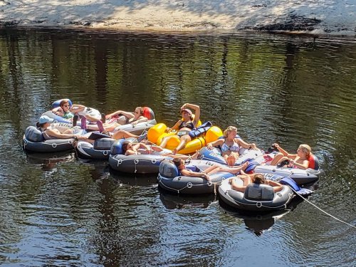 A group of people relaxing on inflatable rafts in a calm river, surrounded by trees and sandy banks.