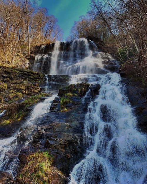A cascading waterfall flows down rocky terrain, surrounded by trees and a clear blue sky.