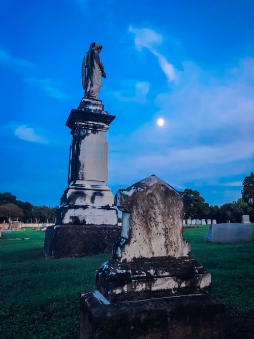 A cemetery scene at dusk featuring a tall angel statue and a weathered gravestone under a bright moon.
