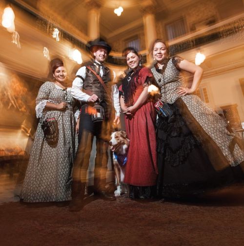A group of four people in vintage attire, posing with a dog in a historic setting, with a blurred background.