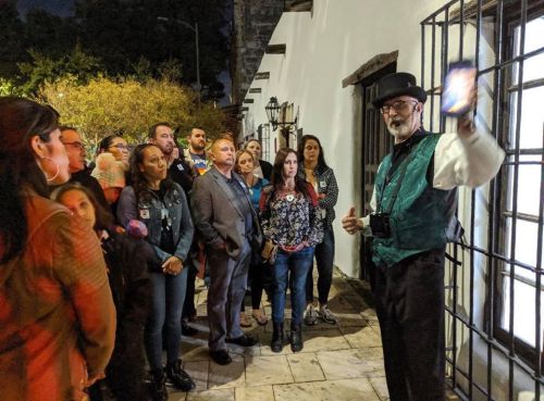 A tour guide in a vest speaks to a captivated group of people outside a historic building at night.
