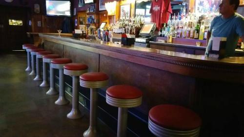 A bar with a polished wooden counter and red cushioned stools, illuminated by warm lighting and a TV in the background.