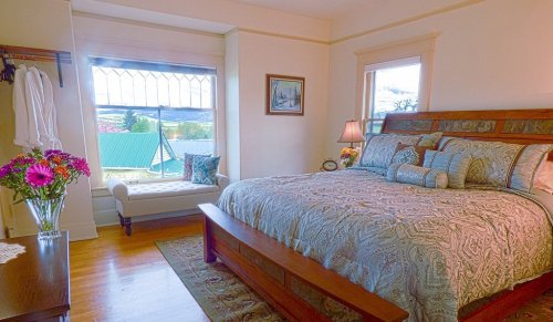 Cozy bedroom with a large bed, decorative pillows, a window seat, and a view of the outdoors. Flowers on a side table.