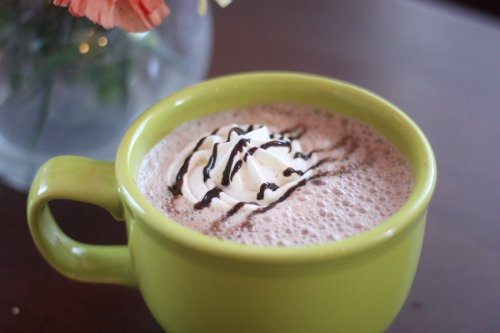 A green mug filled with hot chocolate topped with whipped cream and chocolate drizzle, placed on a wooden table.