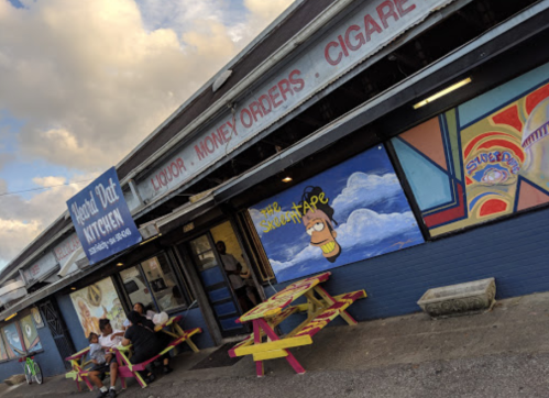 A colorful storefront with a sign for a liquor store and a kitchen, featuring outdoor seating and vibrant murals.