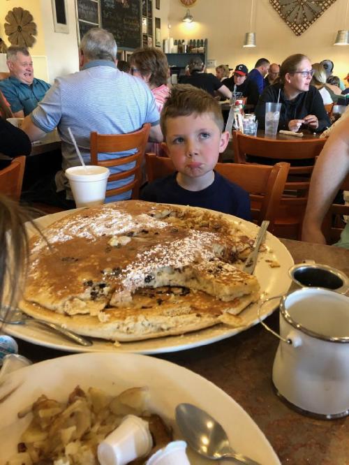 A young boy looks curiously at a giant pancake on a table in a busy restaurant filled with people.