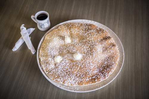 A large, fluffy pancake topped with butter and powdered sugar, served with a small cup of coffee on the side.