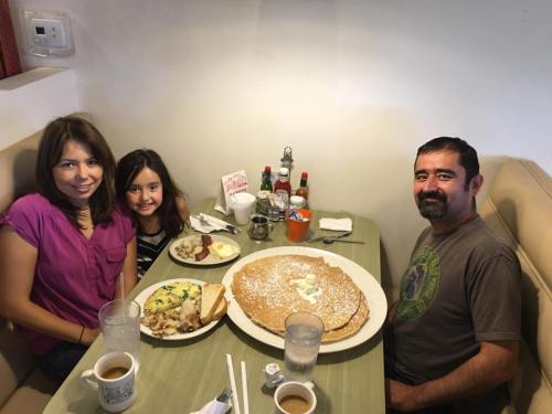 A family of three enjoys a large breakfast with pancakes, eggs, and drinks at a cozy restaurant table.