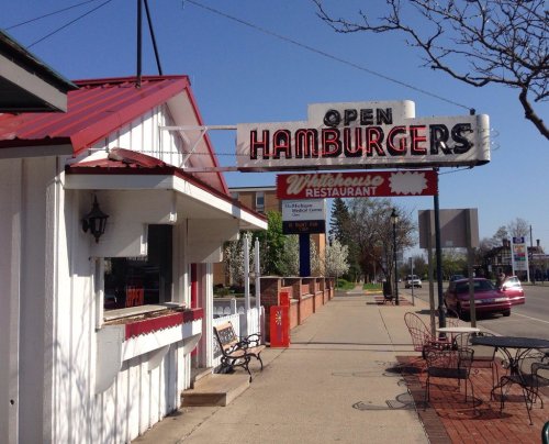 A diner with a red roof and a sign reading "Open Hamburgers" next to a restaurant sign on a sunny street.