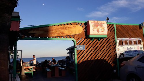 A beachside snack shack with a large surfboard sign, ocean view, and people enjoying the outdoor seating.