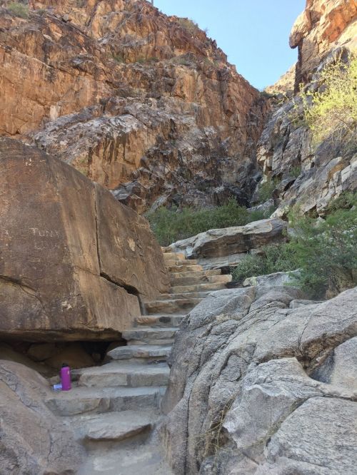 Stone steps lead up a rocky path surrounded by steep cliffs and sparse vegetation under a clear blue sky.