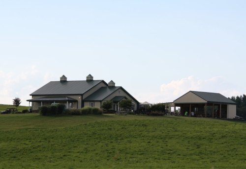 A large, modern farmhouse with a green roof, surrounded by grassy fields and a nearby shed.