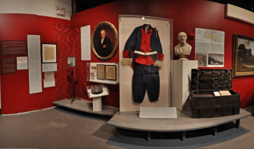 Exhibit featuring historical artifacts, including a uniform, documents, and a bust, set against a red backdrop.