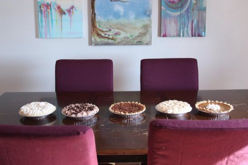 A dining table with five pies of various flavors, flanked by two maroon chairs and colorful artwork on the wall.
