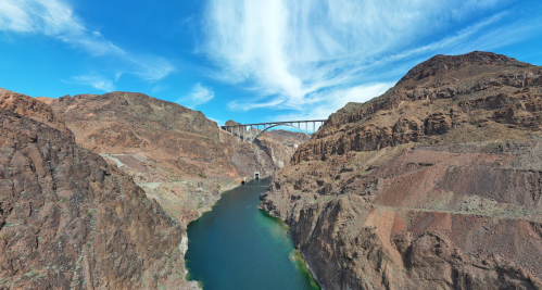 A panoramic view of a river surrounded by rocky cliffs, with a bridge spanning across the landscape under a blue sky.