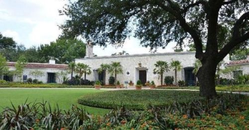 A stone house surrounded by lush greenery and colorful flowers, with large trees in the foreground.