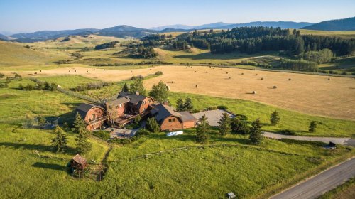 Aerial view of a large rustic house surrounded by green fields and mountains under a clear blue sky.