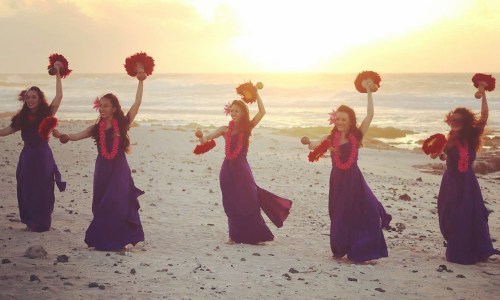 Five dancers in purple dresses and floral leis perform on a beach at sunset, holding maracas and smiling.