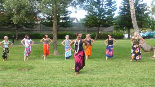A group of women in colorful skirts dancing outdoors on a grassy area surrounded by trees.