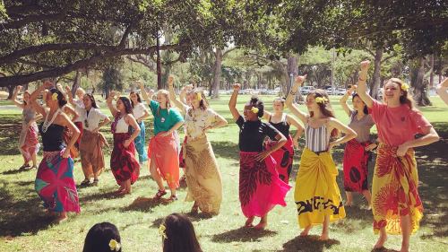 A group of people in colorful skirts dancing outdoors, surrounded by trees and grass, with flowers in their hair.