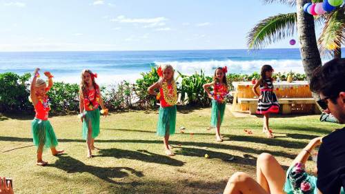 Five children in colorful outfits dance on a beach lawn, with ocean waves and palm trees in the background.