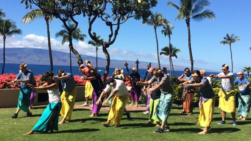 Group of people in colorful skirts dancing outdoors with palm trees and ocean in the background.