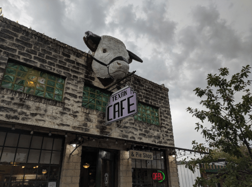 A large cow head sculpture atop a rustic building with a sign reading "Texan Cafe" and an open neon sign.