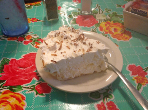 A slice of creamy dessert topped with whipped cream and chocolate shavings on a floral tablecloth.
