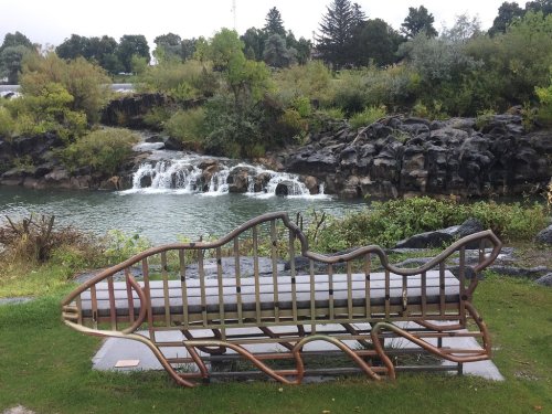 A fish-shaped bench sits near a rocky stream with small waterfalls and lush greenery in the background.