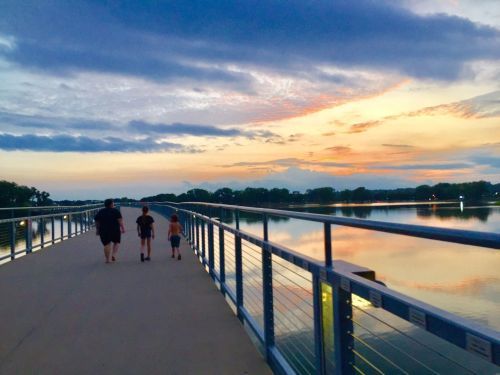 A family walks along a pier at sunset, with calm water reflecting the colorful sky.